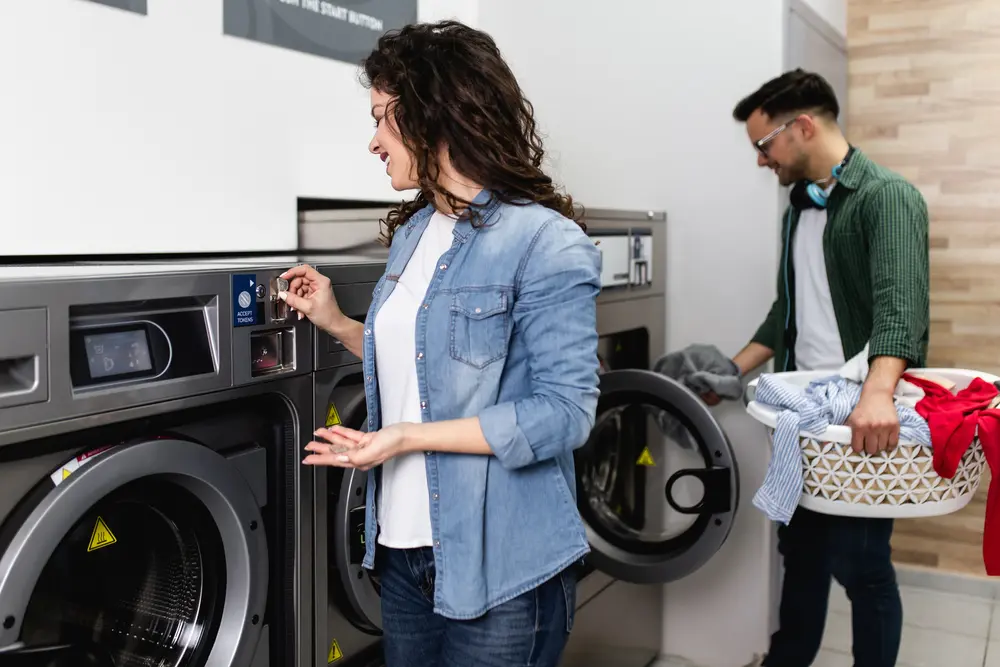Two customers in a laundromat setting holding laundry baskets near commercial washing machines
