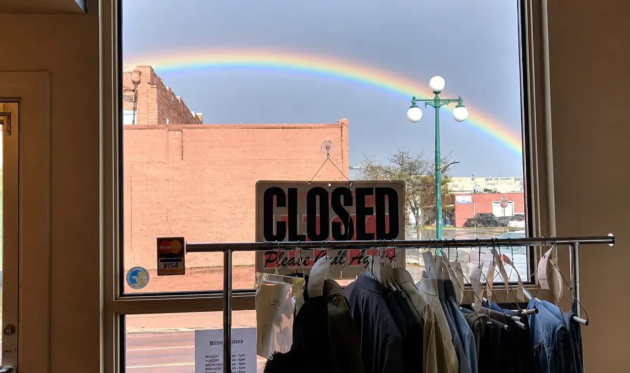 View through a store window with a Closed sign looking out towards street lamp and a bright rainbow in the sky